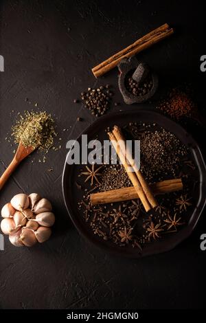 Auswahl an verschiedenen Gewürzen auf texturiertem schwarzem Hintergrund. Pfeffer, Kardamom, Nelke, Knoblauch, Zimt, Star anis, dehydrierte Petersilie. Flach liegend Stockfoto