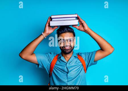 indischer Student Mann glücklich beim Betrachten der Kamera Tragen Sie eine Augenbrille und einen orangen Rucksack im blauen Studio Stockfoto