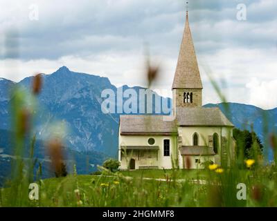 Eine Sommerwiese mit blauem Himmel und ein paar weißen Wolken mit einer großen Bergkette im Hintergrund. Eine kleine Kapelle mit einem knarrigen Obstbaum kann sein Stockfoto