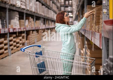 Nowosibirsk, Russland - 02.26.2020: Schöne junge rothaarige Frau beim Einkaufen in Ikea. Porträt eines kaukasischen lächelnden Mädchen mit einem Trolley in einem Stockfoto