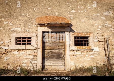 Old Stone Cowshed mit Holztür und Fenstern mit rostigen Eisenstangen. Lessinia Plateau, Bosco Chiesanuova, Provinz Verona, Venetien, Italien. Stockfoto