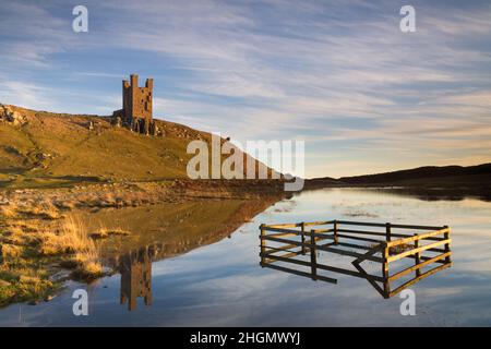 Die Ruinen von Lilburn Tower, Dunstanburgh Castle an der Küste von Northumberland, spiegeln sich in den ruhigen stillen Gewässern eines Sees bei Sonnenuntergang im Winter wider Stockfoto