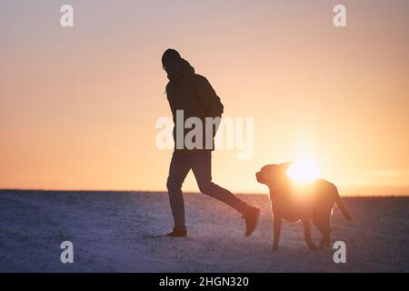 Silhouette eines Mannes mit Hund auf einer verschneiten Wiese. Haustierbesitzer läuft mit labrador Retriever während des Wintermorgens. Stockfoto