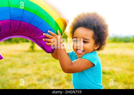 Positive gemischte Rennen kleines Mädchen mit afro lockiges Haar hält Regenbogenballon im Sommerpark Stockfoto