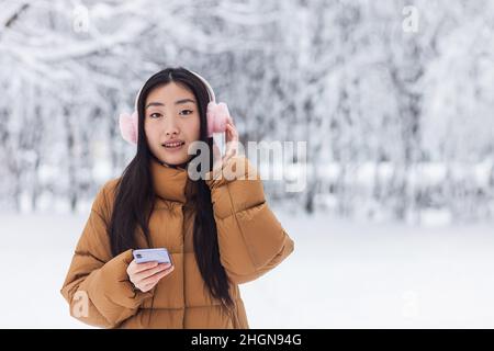 Schöne japanische Teenager geht im Winter Park nutzt das Telefon und hört Musik mit niedlichen rosa Kopfhörer Stockfoto