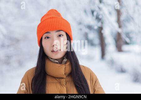 Porträt einer jungen, schönen asiatischen Frau, Nahaufnahme, die in einem winterverschneiten Park die Kamera in warmen Kleidern anschaut Stockfoto