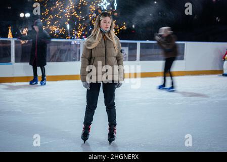 Ein blondes Mädchen im Teenageralter, das auf der Eisbahn mit Weihnachtslichtern im Hintergrund Schlittschuh läuft Stockfoto