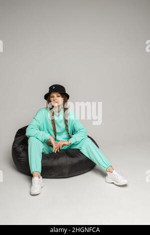 Cooles Mädchen in Hut und sportlicher Anzug sitzt in Tasche Stuhl. Stockfoto