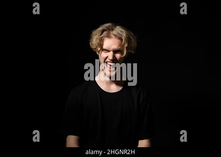 Junger brutaler wütender Mann im schwarzen T-Shirt schreit auf schwarzem Hintergrund. Gesichtsausdruck. Verrückter Mann. Negative Emotionen, Probleme. Gestresster Kerl, der schrie. Stockfoto