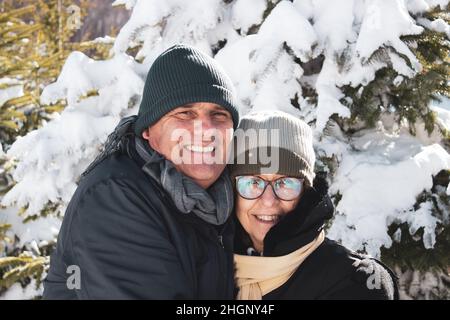 Ehepaar mittleren Alters in Winterkleidung umarmt sich gegenseitig und zeigt Zuneigung. Kaukasisches Paar lächelt zusammen zwischen Schnee und Wald. Candi Stockfoto