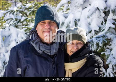 Ehepaar mittleren Alters in Winterkleidung umarmt sich gegenseitig und zeigt Zuneigung. Kaukasisches Paar lächelt zusammen zwischen Schnee und Wald. Candi Stockfoto