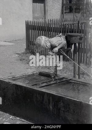 1950s, historisch, Sommerzeit und an einer Dorfwasserpumpe und einem alten Steintrog, ein junges französisches Mädchen, das frisches Wasser aus dem Wasserhahn, Domremy, Frankreich, bekommt. Stockfoto