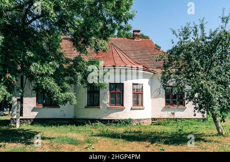 Leeres altes verlassene Gebäude unter großen grünen Bäumen. Dachziegel, Ziegelkamin, Holzfenster. Vintage-Design Stockfoto