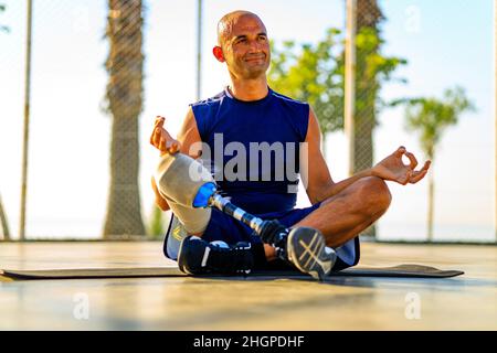 Mann mit Prothesenbein praktiziert Yoga sitzend in Lotushaltung auf Yogamatte, meditierend im Freien in Sonnenlichtern Stockfoto
