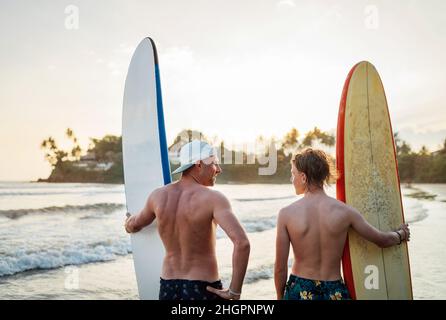 Vater mit Teenager-Sohn, der mit Surfbrettern am sandigen Meeresstrand mit Palmen im Hintergrund steht, die von der Sonne des Sonnenuntergangs erhellt werden. Sie lächeln und h Stockfoto