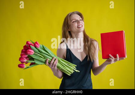 Frau mit einem Strauß roter Tulpen auf gelbem Hintergrund. Glückliches Mädchen in einem schwarzen Kleid hält eine Armvoll Blumen und eine Schachtel Pralinen. Geschenk für Stockfoto