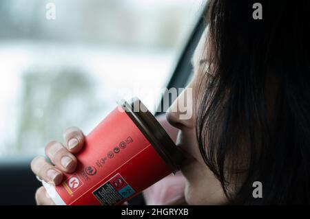 Frau mit schwarzen Haaren sitzt im Auto und trinkt Kaffee aus einer recycelbaren Papiertasse Stockfoto