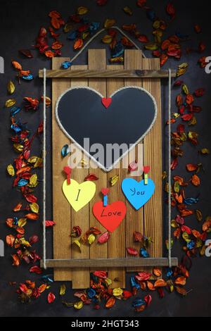 Leere Herztafel auf Holz. Valentinstag-Konzept. Dunkler Hintergrund mit bunten trockenen Rosenblättern. Herzförmige Notizpapiere mit „I Love You“ wri Stockfoto