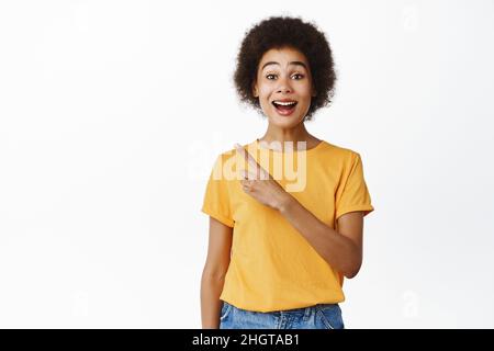 Enthusiastische schwarze Frau, die mit dem Finger auf die linke obere Ecke zeigt und lächelt, neugierig auf das Promo-Angebot, überrascht vom Verkauf, zeigt das Firmenlogo zur Seite Stockfoto