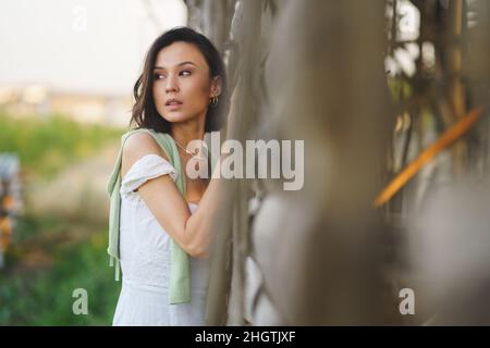 Asiatische Frau, die in der Nähe eines Tabaktrocknungsschuppens posiert und ein weißes Kleid und grüne Gummistiefel trägt. Stockfoto