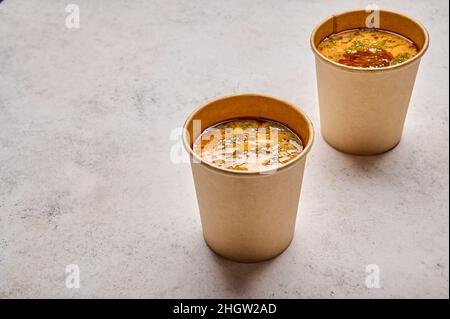Nahaufnahme Tom Yam Suppe mit Lachs und Meeresfrüchten in zwei festen Papierbehältern auf hellem Hintergrund selektiver Fokus. Designer-Food-Konzept für die Mitnahme oder Lieferung Stockfoto