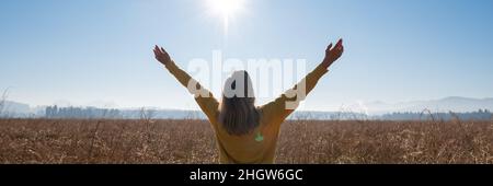 Breitbild-Bild einer jungen Frau in gelbem Pullover, die an einem schönen sonnigen Tag auf einem Feld mit weit ausgebreiteten Armen steht. Stockfoto