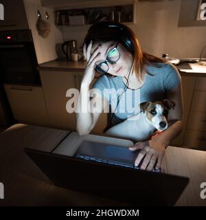 Müde Frau in Kopfhörern und Brillen, die Überstunden über einem Laptop machen. Freiberufliche Mitarbeiterin, die spät am Abend zu Hause in der Küche aus der Ferne arbeitet Stockfoto