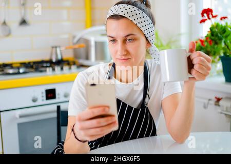 Frau sitzt am Tisch und nutzt Smartphone für Nachrichten zu sehen, Chat während in der Küche entspannen . Konzept der Entspannung Stockfoto