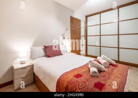 Schlafzimmer mit eingebautem Kleiderschrank mit holzgerahmten, rötlich dekorierten Schiebetüren aus Glas, Rollhandtüchern und hellen Nachttischen Stockfoto