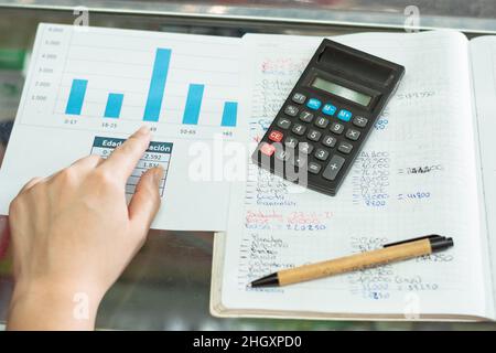 Draufsicht auf die Hand eines Mädchens, die auf ihr Geschäftsstatistikdiagramm zeigt. Neben ihr ein umweltfreundlicher Pappstift und ein kleiner schwarzer Rechner, oben Stockfoto