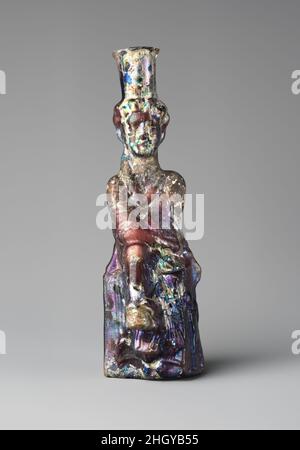 Glasflasche in Form von Tyche (Good Fortune) 2nd–3rd century A.D. römisch-lichtdurchlässiges Purpur.Evertierter, unebener Rand, in der Flamme abgerundet; zylindrischer Hals, leicht nach unten zugeneigt; länglicher Körper; eingeschubter Boden mit Kreis in geringem Relief. Eine durchgehende Schimmelnaht, leicht verstellt, die vom Rand um den Körper und über den Boden läuft.Körper, der durch Schimmel in Form einer weiblichen Figur geformt wurde, wobei das Haar in ein Brötchen zurückgezogen wurde, drapiert, die Arme gefaltet, auf einem polygonalen Sockel oder Stuhl sitzend, richtiger rechter Fuß auf stilisiertem Kopf ruhend; Auf der rechten linken Seite des Sockels im Relief, stehend geflügelte Eros Stockfoto