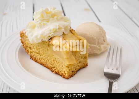 Stück Apfelkuchen mit Biskuitboden und Schlagsahne Belag, serviert mit Vanilleeis aus nächster Nähe auf einem Teller auf weißem Holzhintergrund Stockfoto