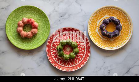 Drei bunte Teller mit geometrischem Design, mit drei verschiedenen Mochi Donuts. Erdbeer-, Ube- und Matcha-Glasur. Stockfoto