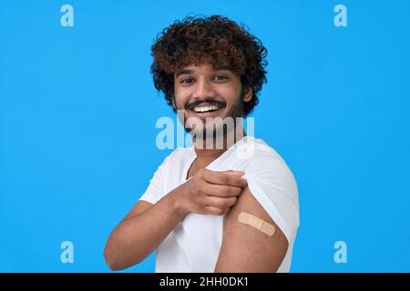 Glücklicher junger indischer Mann, der Verband am Arm zeigt, nachdem er die Impfung auf Blau isoliert hatte. Stockfoto