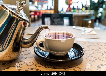 Der Typ hält den Wasserkocher und gießt heißen Tee in die Tasse. Cafe. Roter heißer Kräutertee im Restaurant. Tee gießen Stockfoto