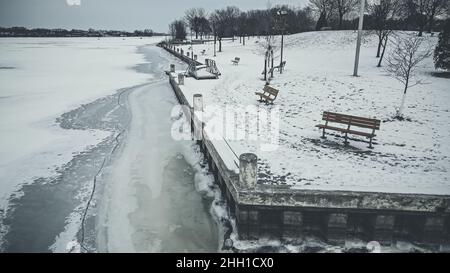 Die Küste ist gefroren und die Läufe entlang des Parks. Schnee bedeckt das Land mit Bäumen und Bänken. Stockfoto