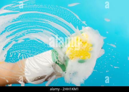 Weibliche Hand in Gummihandschuhen wischt das Glas mit einem gelben Schaumschwamm ab. Hellblauer Hintergrund. Das Konzept des professionellen Reinigungsservice. Stockfoto
