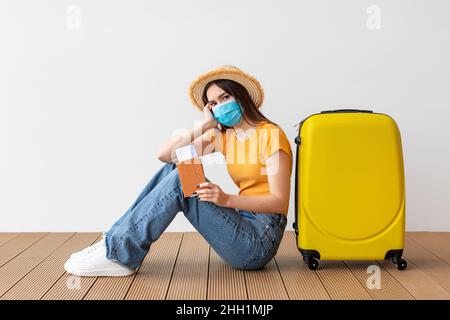 Verärgerte Frau, die eine schützende medizinische Maske trägt, ihren Pass mit Ticket und Bordkarte in der Nähe ihres Gepäcks hält Stockfoto