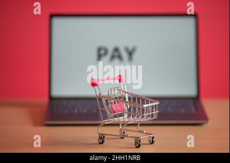 Mini-Einkaufswagen steht vor einem Laptop auf einem rosa Hintergrund. Online-Shopping-Konzept. Das Wort Pay auf einem Computerbildschirm. Remote-Kauf von Stockfoto