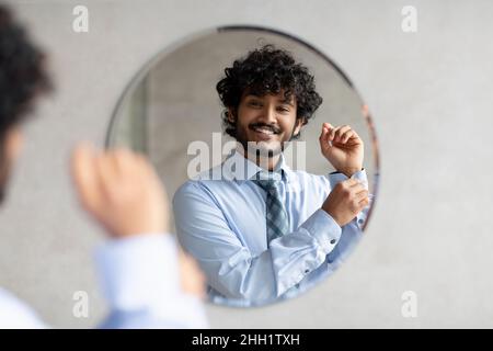 Glücklicher bärtiger indischer Geschäftsmann, der im Badezimmer vor dem Spiegel stand und ein Hemd aufknöpfte Stockfoto