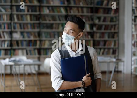 Nachdenklicher asiatischer College-Kerl in Maske besucht Universitätscampus-Bibliothek Stockfoto