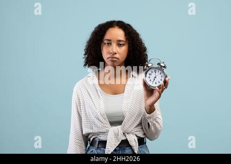 Gelangweilte junge schwarze Studentin, die Wecker vor der Kamera über blauem Studiohintergrund zeigt Stockfoto
