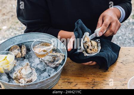 Frau schüttelt Austern am Tisch im Freien mit einem mit Eis gefüllten Behälter mit Austern und Gewürzen Stockfoto