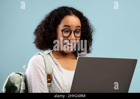 Emotional junge afroamerikanische Frau mit Rucksack Blick auf Laptop-Bildschirm in Aufregung über blauen Hintergrund Stockfoto