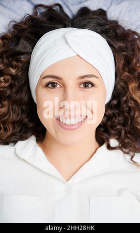 Draufsicht auf eine schöne junge Frau mit lockigen Haaren und weißem Stirnband, während sie die Kamera ansah und lächelte. Charmante weibliche Person mit perfekter Haut, die positive Emotionen ausdrückt. Stockfoto