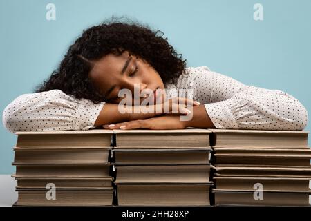 Müde afroamerikanische Studentin, die auf Bücherstapeln schläft und während der Vorbereitung auf die Prüfungen erschöpft ist Stockfoto