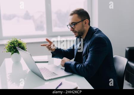 Fokussierter junger Geschäftsmann in einer Brille mit Kopfhörern, der Videoanruf mit Kunden auf dem Laptop hielt. Stockfoto