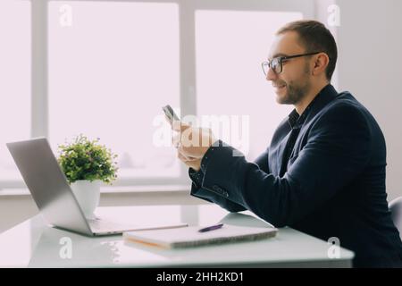 Manager in formeller Kleidung hält Mobiltelefon in den Händen, denkt allein im Büro an Geschäftschancen, lächelt der Vorgesetzte, plant den Arbeitstag, fühlt sich aufgergt Stockfoto