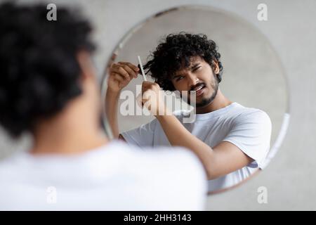 Männliche Schönheitspflege und Haarprobleme. Trauriger indischer Mann im weißen T-Shirt kann die Haare nicht kämmen und hat Schmerzen und Schwierigkeiten Stockfoto