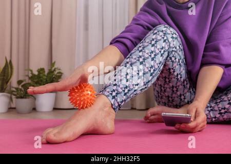 Das Mädchen sitzt auf dem Boden und massiert sich mit einem stacheligen Ball, um sich zu entspannen und zu zirkulieren Stockfoto